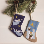 Two embroidered Christmas stockings with nativity scene designs hanging from a branch.