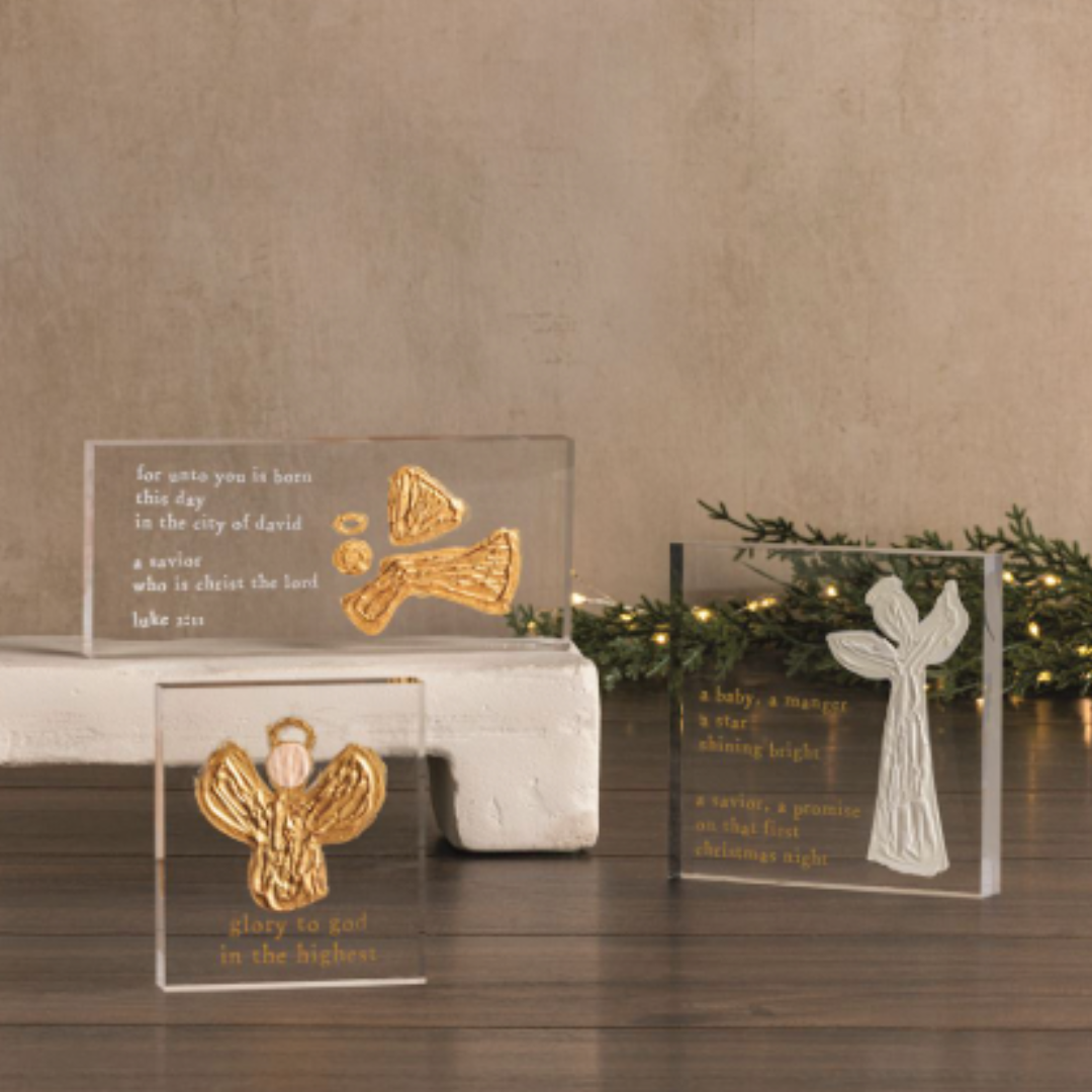 Decorative angel figurines in clear glass cases on a wooden surface with a beige wall and greenery in the background.