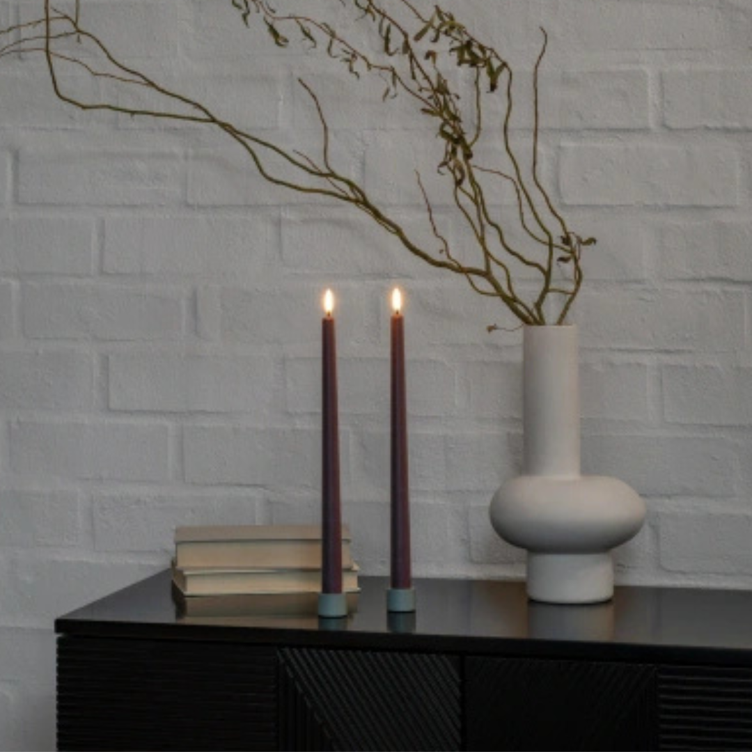 Two lit candles on a table with a vase and books against a white brick wall.