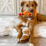 dog holding Plush orange “Furball Cinnamutt Whisky” dog toy shaped like a whiskey bottle with embroidered label and built-in squeaker.