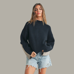 Woman wearing a black sweater with beige accents and denim shorts on a gray background