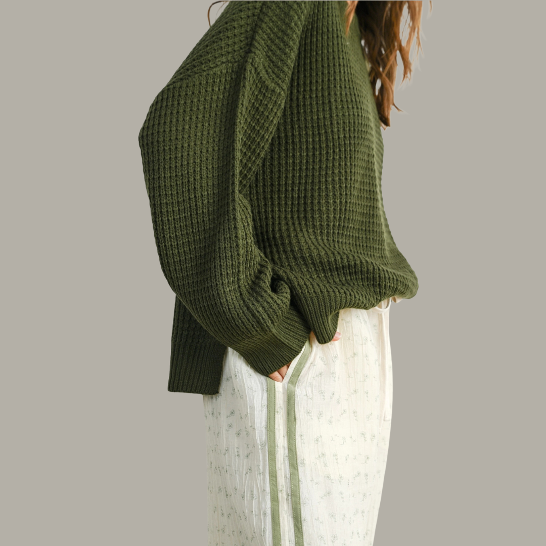 Person wearing a green knitted sweater and white pants on a plain background