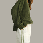 Person wearing a green knitted sweater and white pants on a plain background