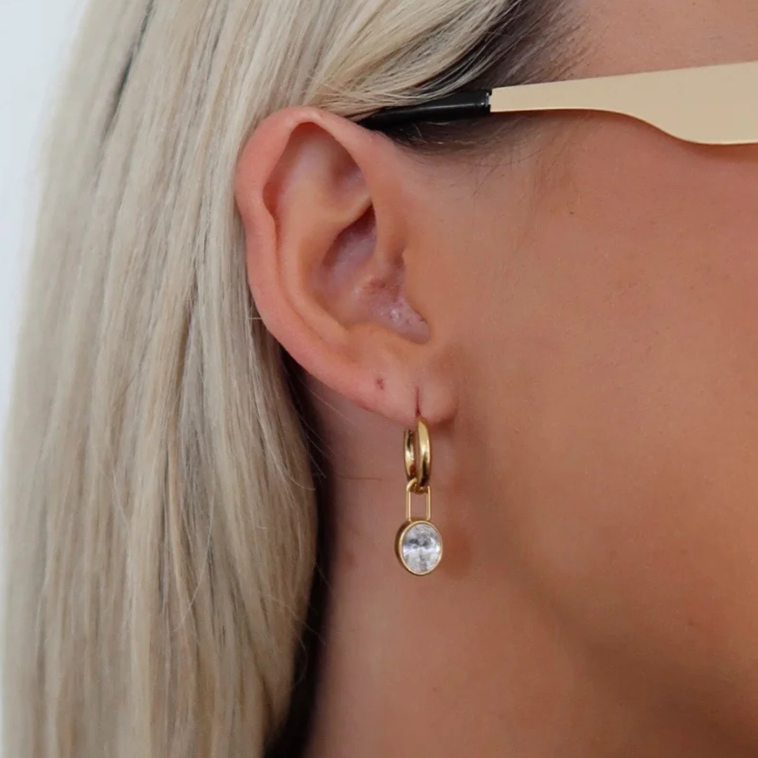 Gold earring with a clear gemstone worn by a person with blonde hair.