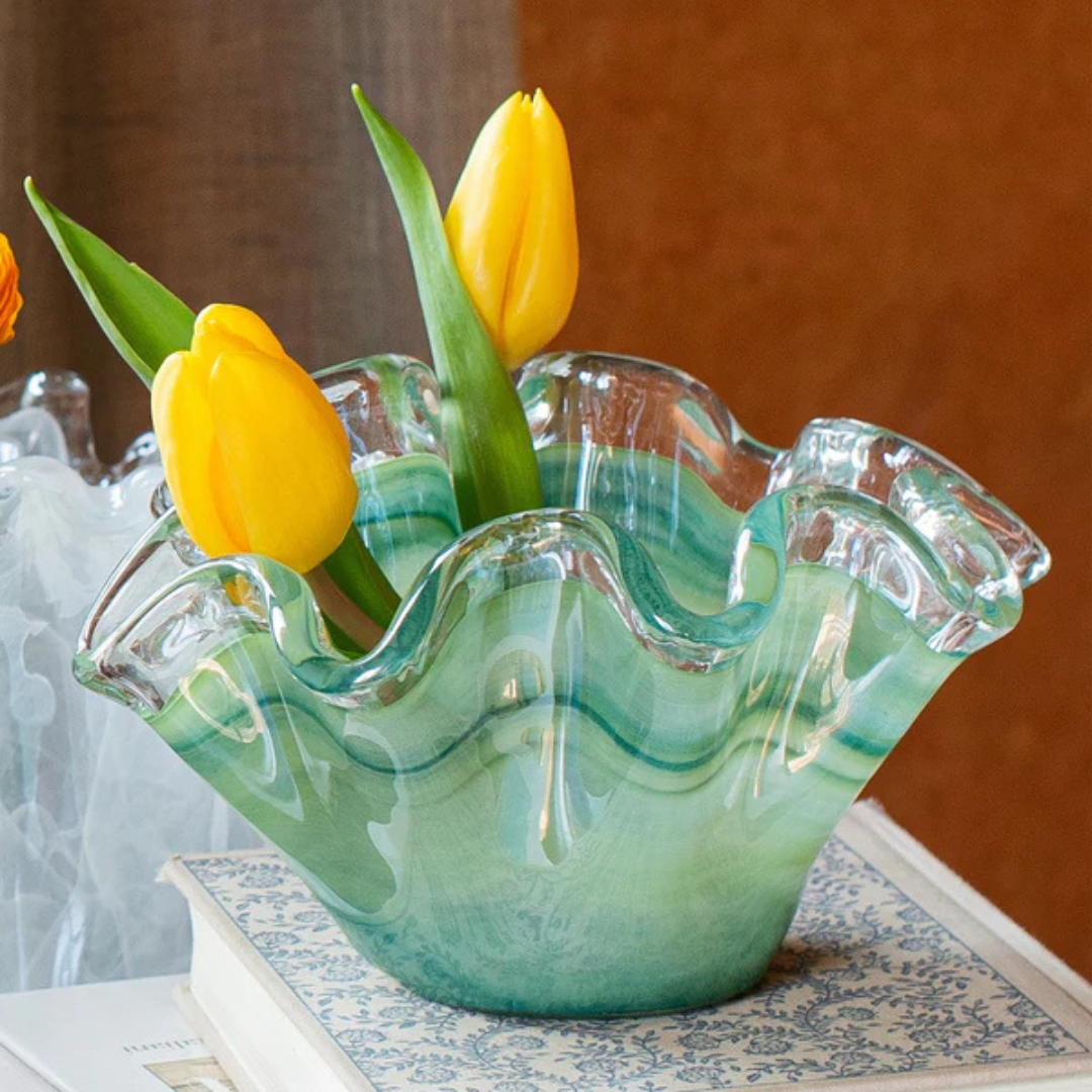 Glass vase with wavy design containing yellow tulips on a patterned surface.