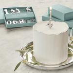 White cake with a lit candle on a silver tray, surrounded by decorative items including a box labeled 'Mariposa'.