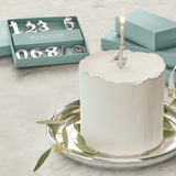 White cake with a lit candle on a silver tray, surrounded by decorative items including a box labeled 'Mariposa'.