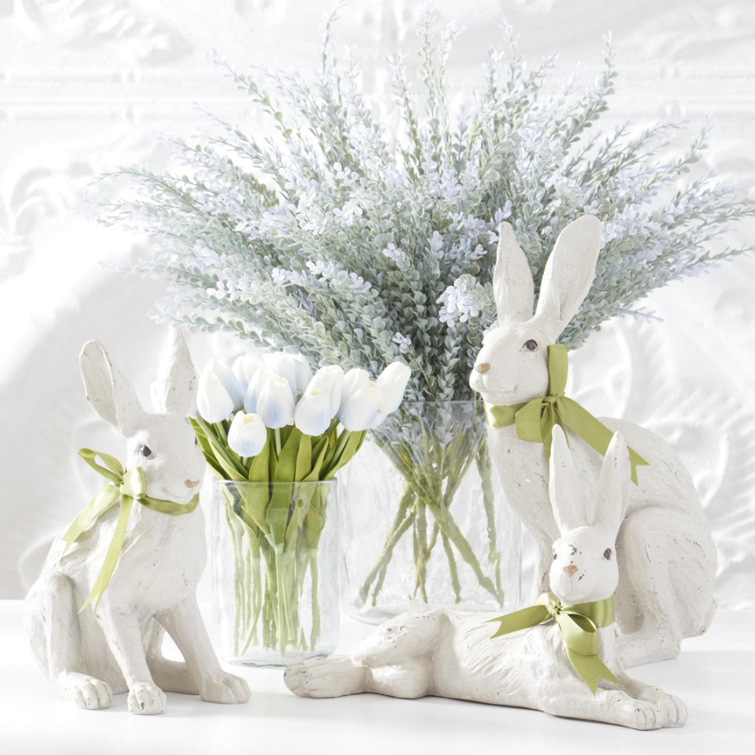 Decorative white rabbits with green ribbons in front of a vase with white tulips and a large floral arrangement on a white background.