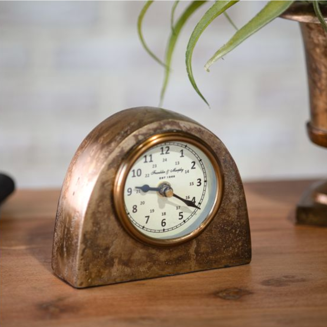 Vintage-style clock on a wooden surface with a blurred plant in the background