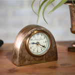 Vintage-style clock on a wooden surface with a blurred plant in the background
