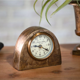 Vintage-style clock on a wooden surface with a blurred plant in the background