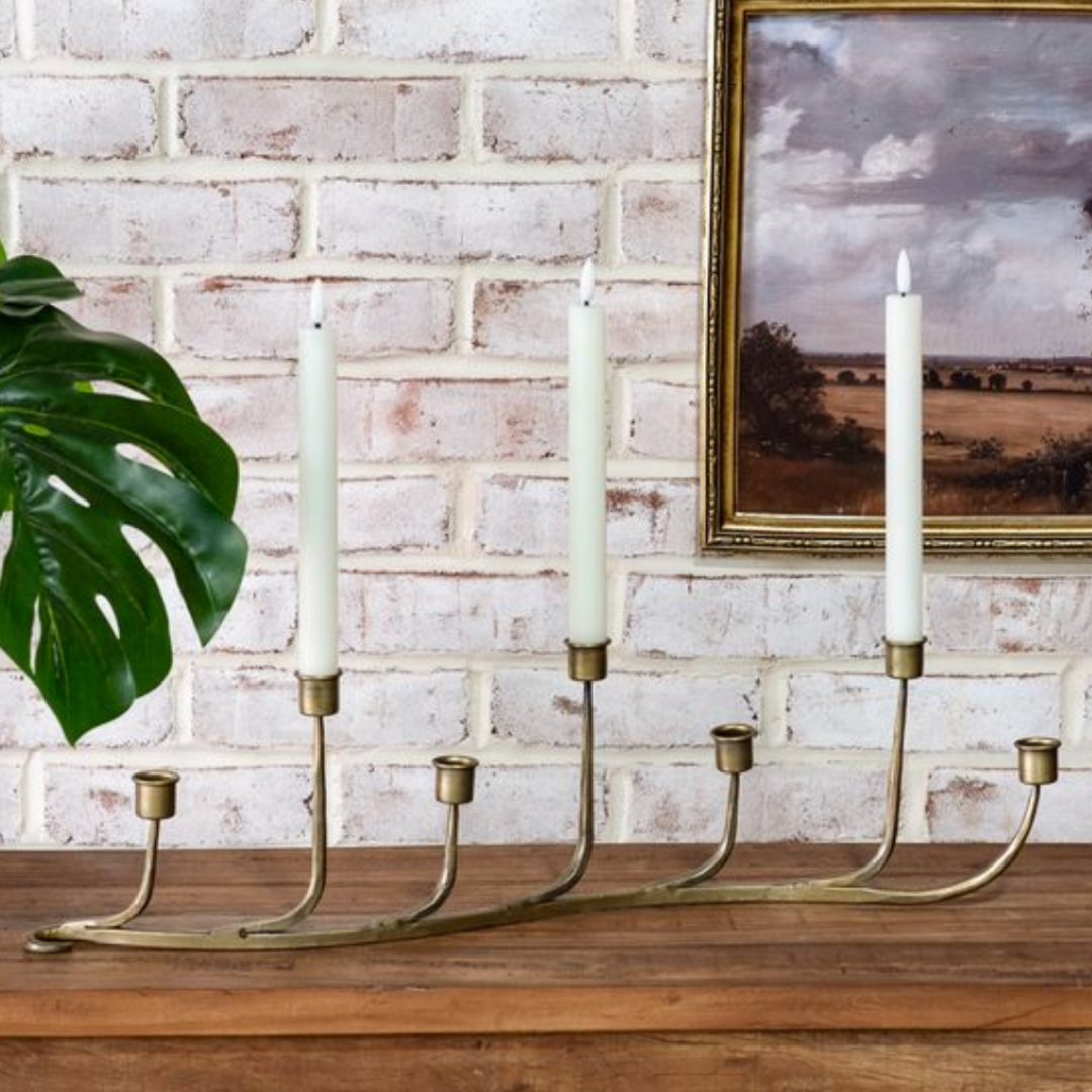 Brass candle holder with three candles on a wooden surface against a brick wall with a painting.