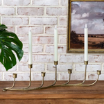 Brass candle holder with three candles on a wooden surface against a brick wall with a painting.