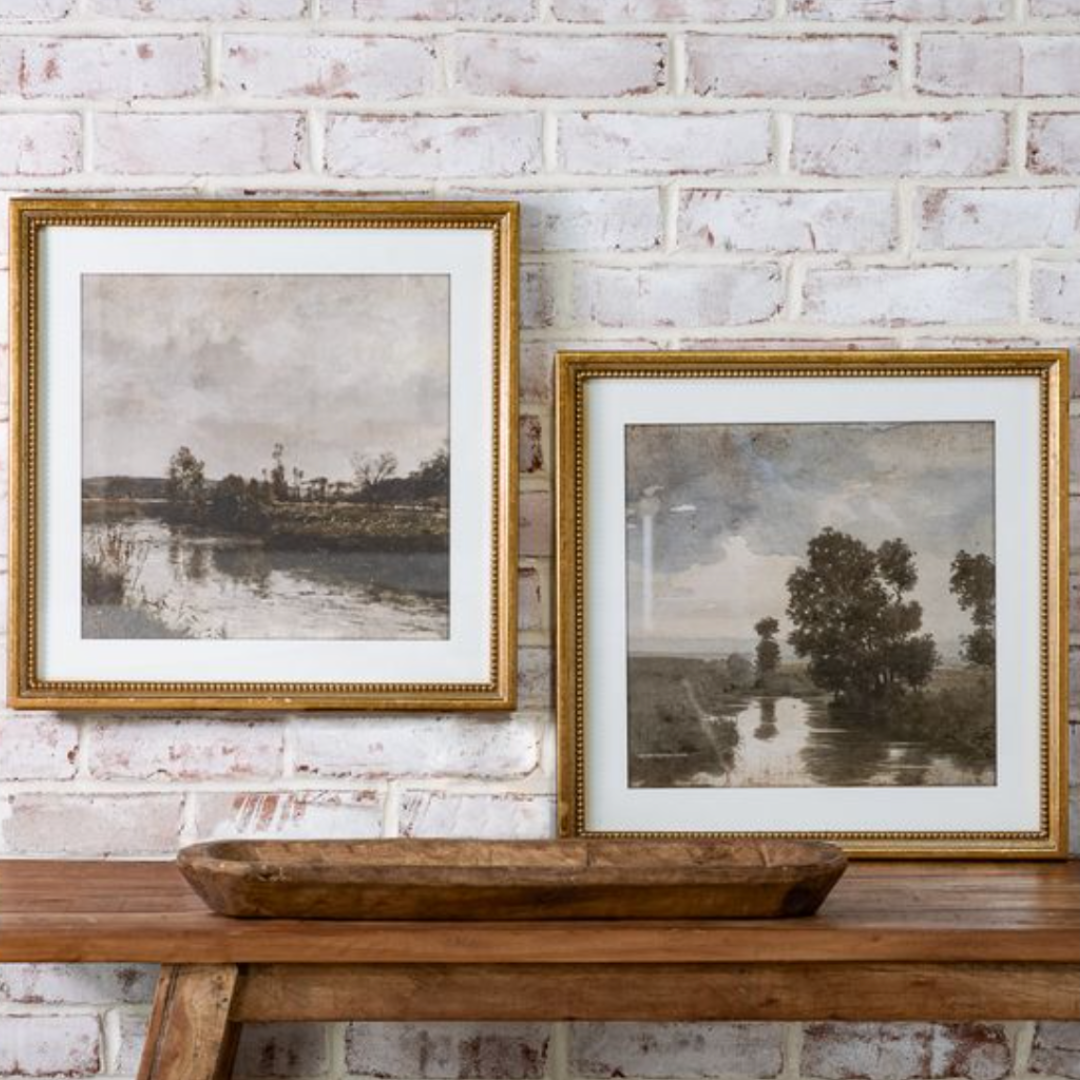 Two framed paintings on a wooden surface against a brick wall.