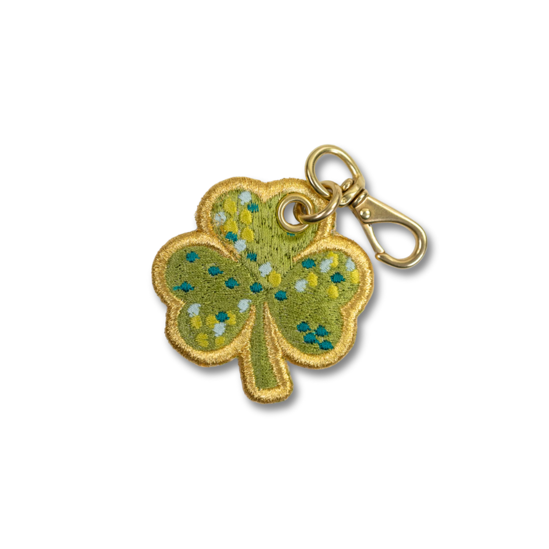 An embroidered shamrock charm with a gold-tone metal clasp, featuring green and yellow embroidered leaves with blue and white floral details.