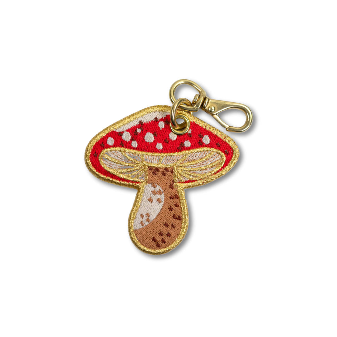 Embroidered mushroom keychain charm with a red cap, white spots, brown stem, and gold-tone metal clasp on a white background.