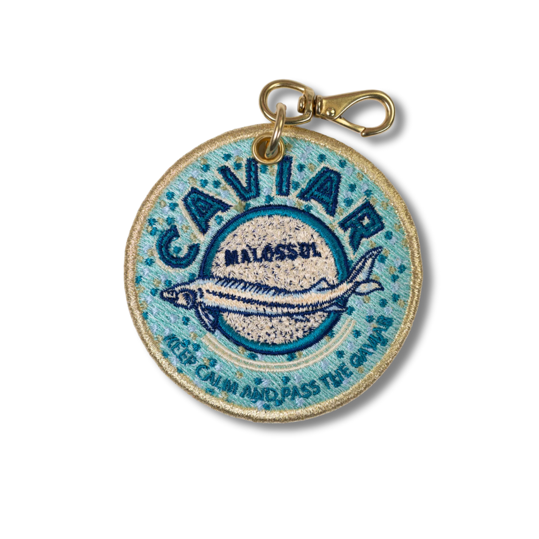 A round embroidered keychain charm featuring a blue fish design and the text 'CAVIAR MALOSSA' with a gold-tone lobster clasp attached at the top, shown on a plain white background.