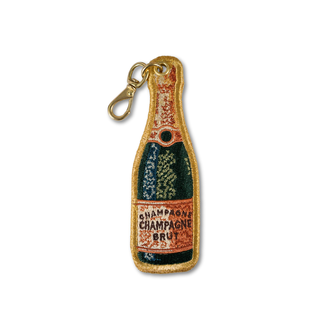 An embroidered keychain charm shaped like a champagne bottle, featuring a gold clasp. The embroidery depicts a dark green bottle body with a distressed orange label reading 'CHAMPAGNE CHAMPAGNE BRUIT' and gold glittery trim.