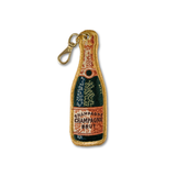 An embroidered keychain charm shaped like a champagne bottle, featuring a gold clasp. The embroidery depicts a dark green bottle body with a distressed orange label reading 'CHAMPAGNE CHAMPAGNE BRUIT' and gold glittery trim.