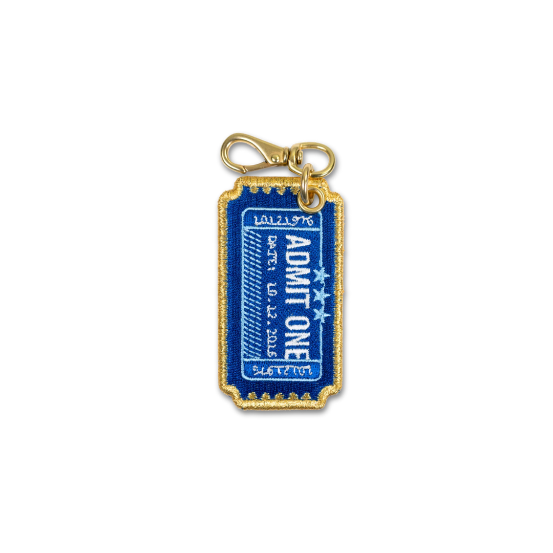 A gold-tone embroidered keychain charm shaped like a blue and white 'ADMIT ONE' ticket, complete with a gold clasp at the top, shown on a plain white background.