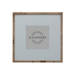 A square picture frame with a natural light brown wood border and a light gray mat, featuring a central gray placeholder card with the brand name 'DANSHIRE' printed in black.