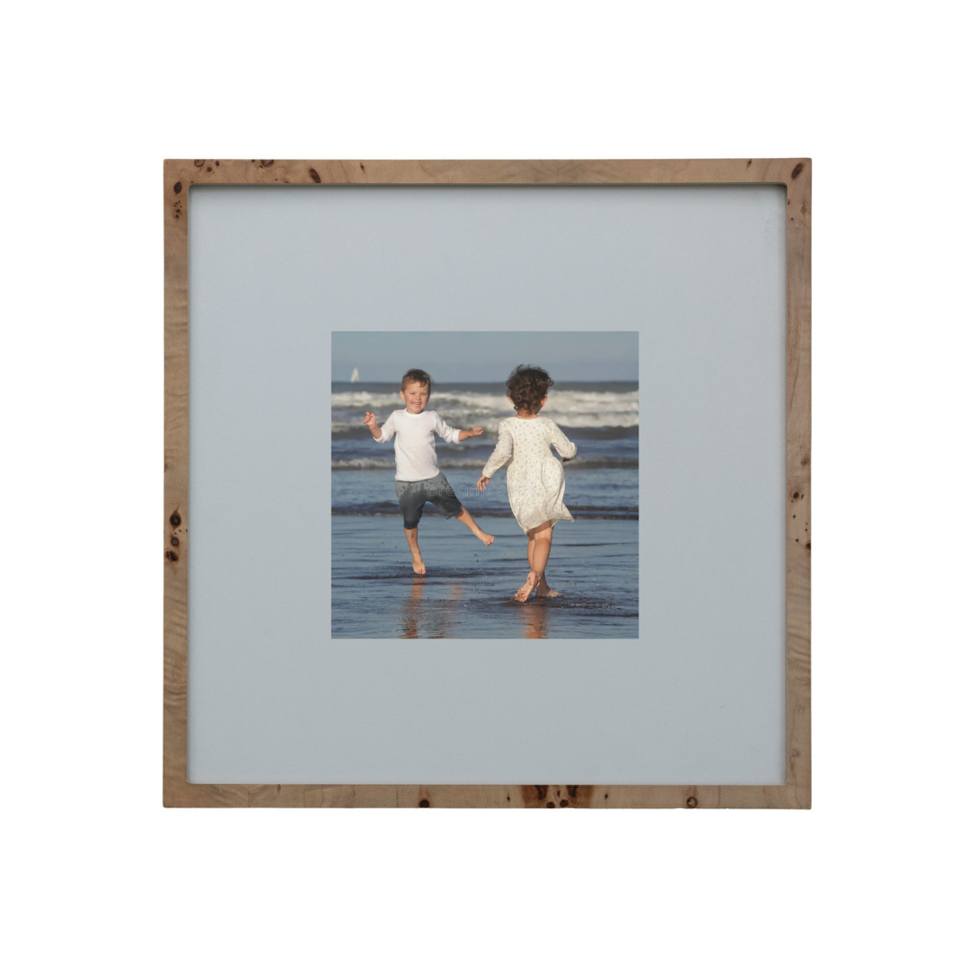 A square wooden picture frame with a light natural wood border and a wide light gray mat, displaying a central photograph of two children running hand in hand on a beach.