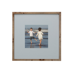 A square wooden picture frame with a light natural wood border and a wide light gray mat, displaying a central photograph of two children running hand in hand on a beach.