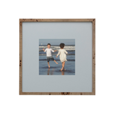 A square wooden picture frame with a light natural wood border and a wide light gray mat, displaying a central photograph of two children running hand in hand on a beach.