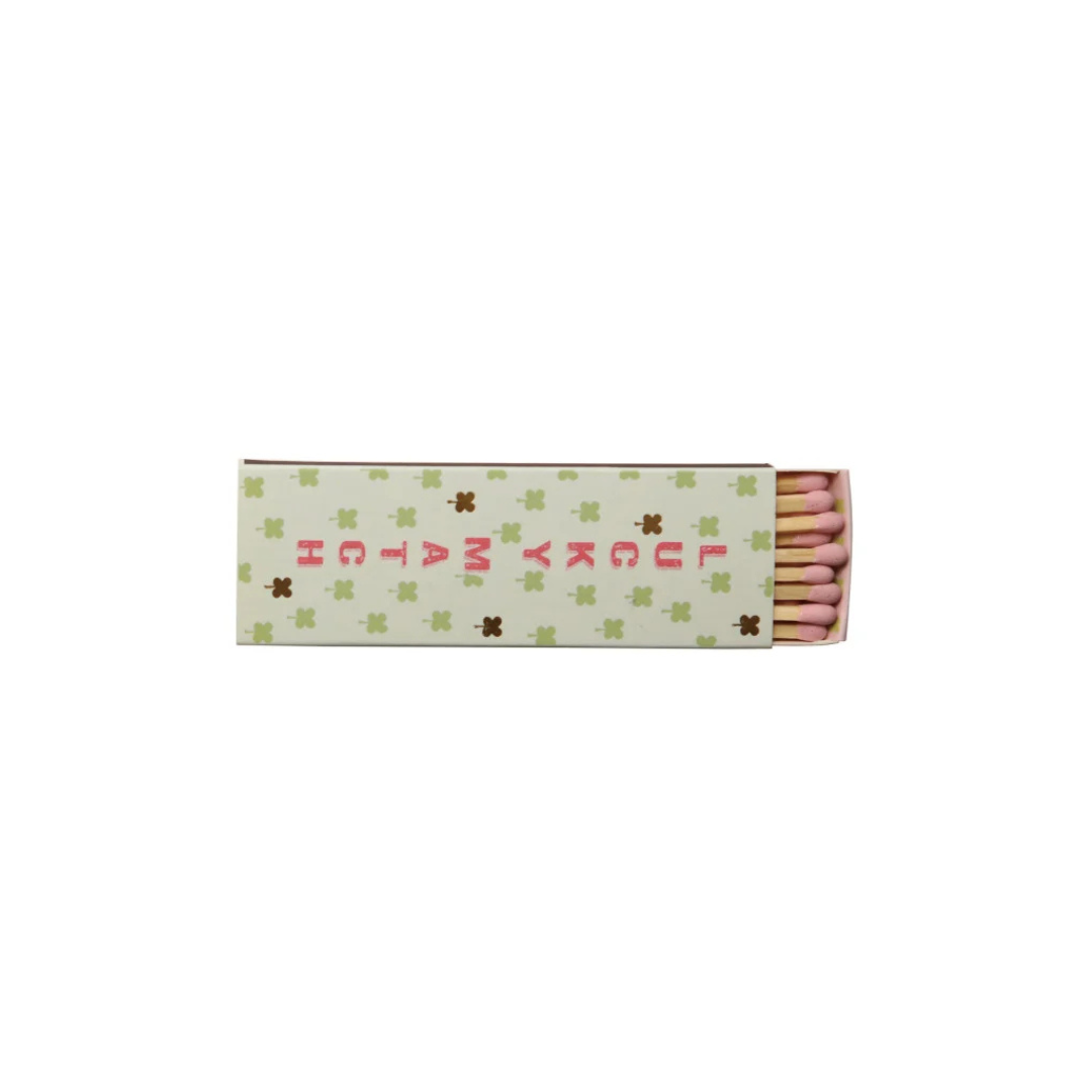 A matchbox with a cream-colored cover decorated with green star patterns and pink text reading 'LUCKY MATCH', with pink matches visible on the right side, set against a plain white background.