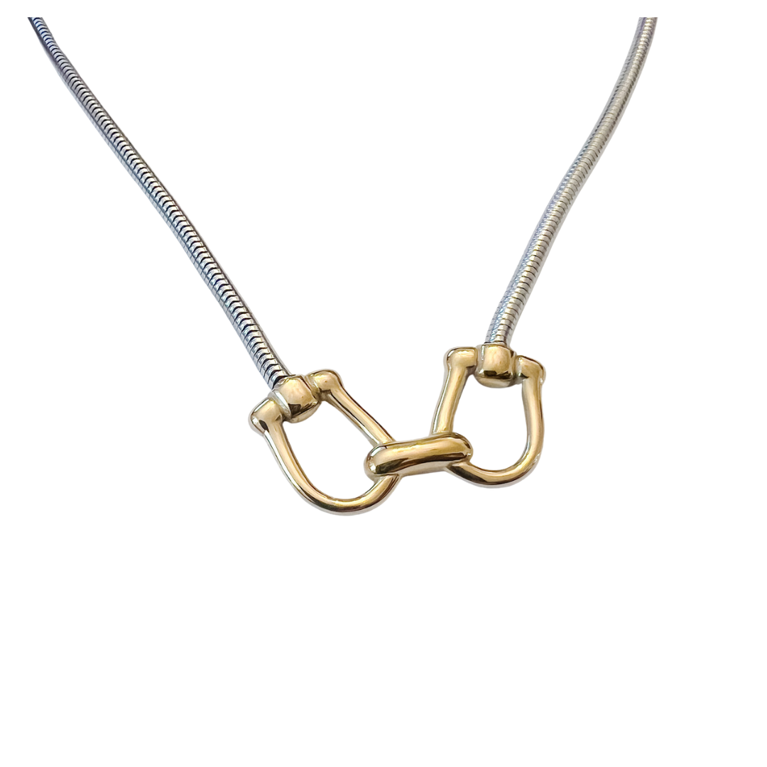 Silver snake chain necklace featuring a gold equestrian horse-bit centerpiece with mixed metal detailing on a white background.