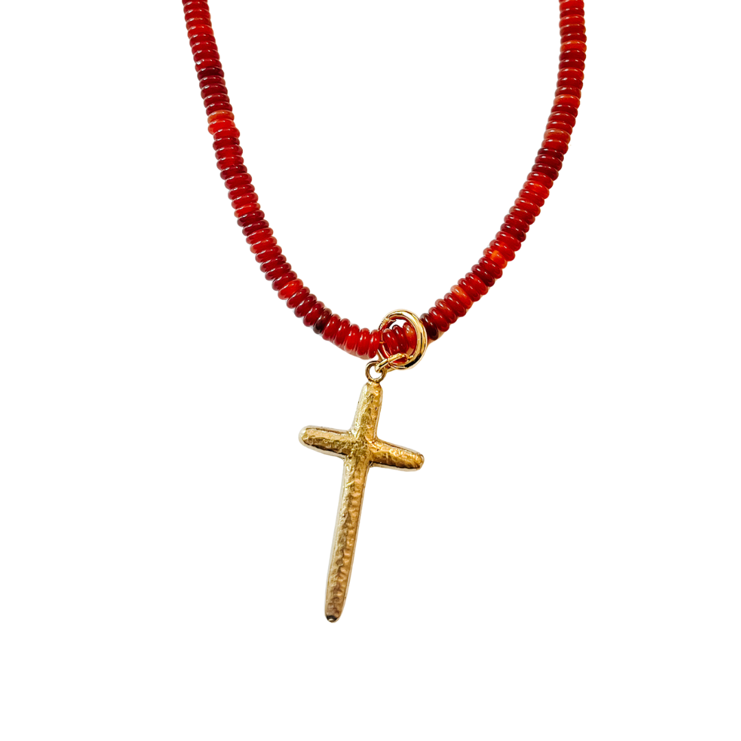 Hammered gold cross charm hanging from a red beaded necklace on a white background.