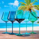 Five colorful wine glasses on a table with a beach and palm tree in the background
