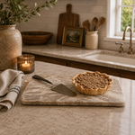 A baked pie with a crumb topping sits on a rectangular stone serving board on a kitchen countertop, with a pie server beside it. The scene includes a vase with greenery, a lit candle, a towel, a cutting board, and a sink in the background.