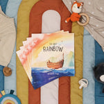 Children's book titled 'The First Rainbow' by Jordan P. Frye on a colorful blanket with toys around.