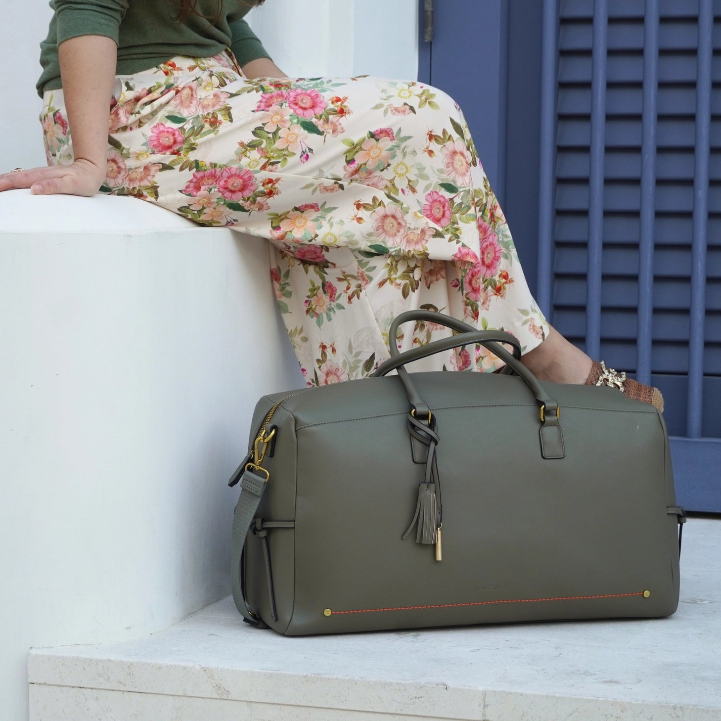 Person sitting on a white bench with a Montemare Roma Duffle in Olive next to them, wearing floral pants.