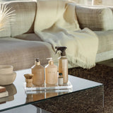 A set of three beige and gold La Prairie home fragrance products on a glass tray, styled on a glass coffee table in a cozy living room setting with a beige sofa and cream throw blanket.