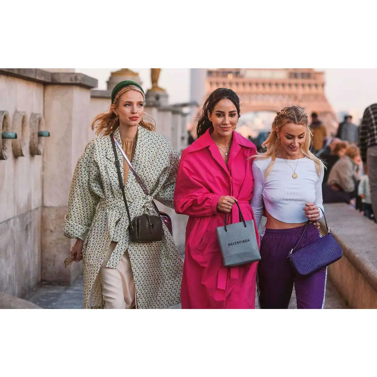 Three women walking together with fashionably styled outfits and handbags in front of the Eiffel Tower.