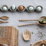 Wooden utensils and a bread loaf on a light gray surface with eggs in a wooden holder.