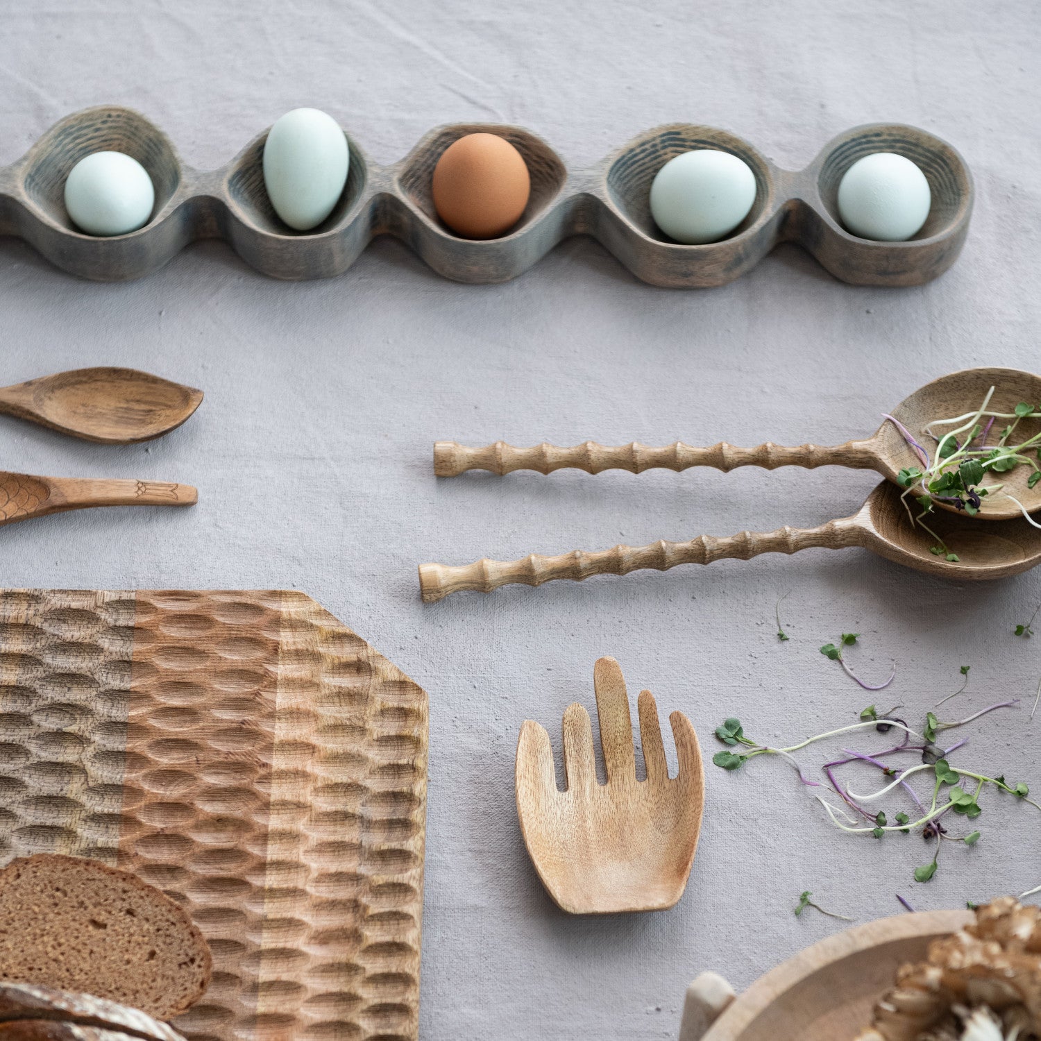 Wooden utensils and a bread loaf on a light gray surface with eggs in a wooden holder.