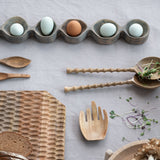 Wooden utensils and a bread loaf on a light gray surface with eggs in a wooden holder.