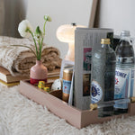 A blush pink wooden tray styled on a white faux fur surface, holding a folded newspaper, a pink vase with white flowers, a can of Smirnoff, a bottle of Stolichnaya, a clear bottle, and a glass. A folded beige blanket and books sit behind it on a soft surface.