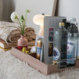 A blush pink wooden tray styled on a white faux fur surface, holding a folded newspaper, a pink vase with white flowers, a can of Smirnoff, a bottle of Stolichnaya, a clear bottle, and a glass. A folded beige blanket and books sit behind it on a soft surface.