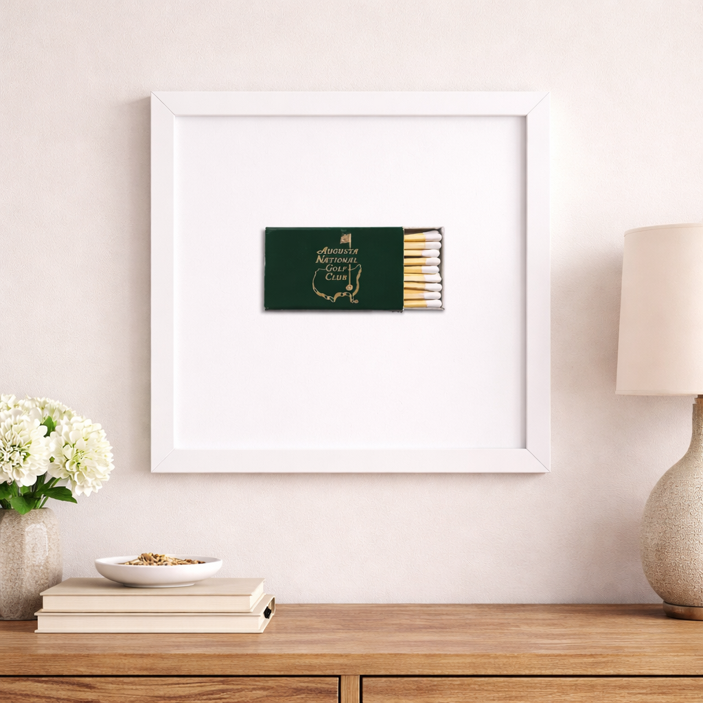 Framed artwork of a matchbox on a wall above a wooden surface with decor items.