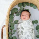 Newborn baby wrapped in a floral blanket lying in a woven basket with green leaves.