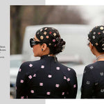 Two women wearing black outfits with floral patterns and Chanel headscarves, with a blurred background.