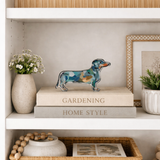 Small acrylic dachshund dog shelfie displayed on stacked neutral books inside a styled white bookcase with a vase of flowers and framed art.