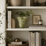 A styled interior shelf scene with books, a ceramic vase, a green marbled planter with leafy stems, framed art, a candle, and a woven tray on a light neutral wall.