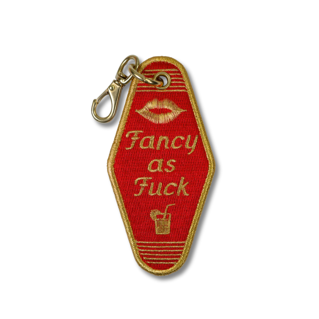A vintage hotel room key embroidered patch in red with gold trim, featuring the text 'Fancy as Fuck' in gold lettering, a gold lip print, and a small cocktail icon. The patch is attached to a gold metal carabiner clip and shown on a plain white background.