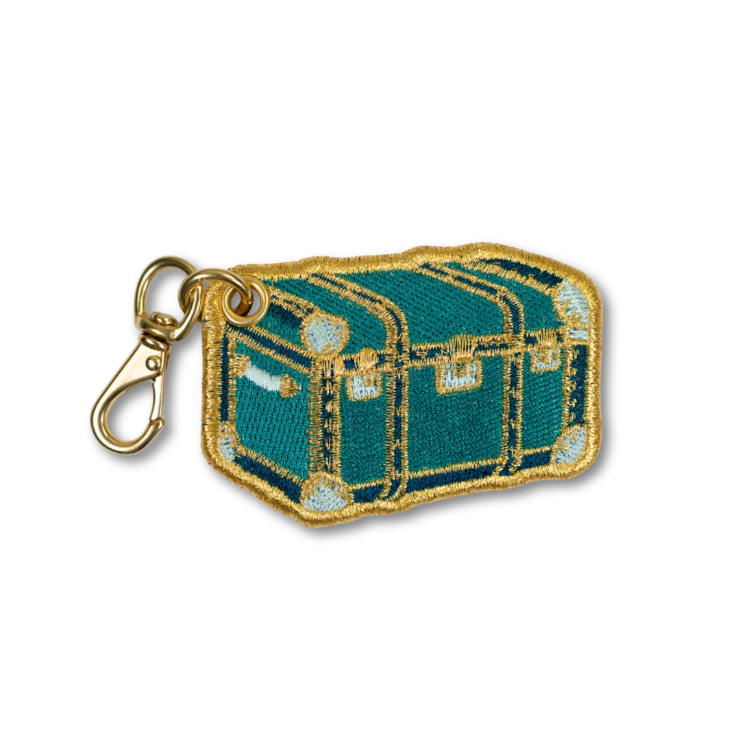 An embroidered patch shaped like a treasure chest in teal green with gold metallic embroidery and a gold lobster clasp attached, shown on a plain white background.