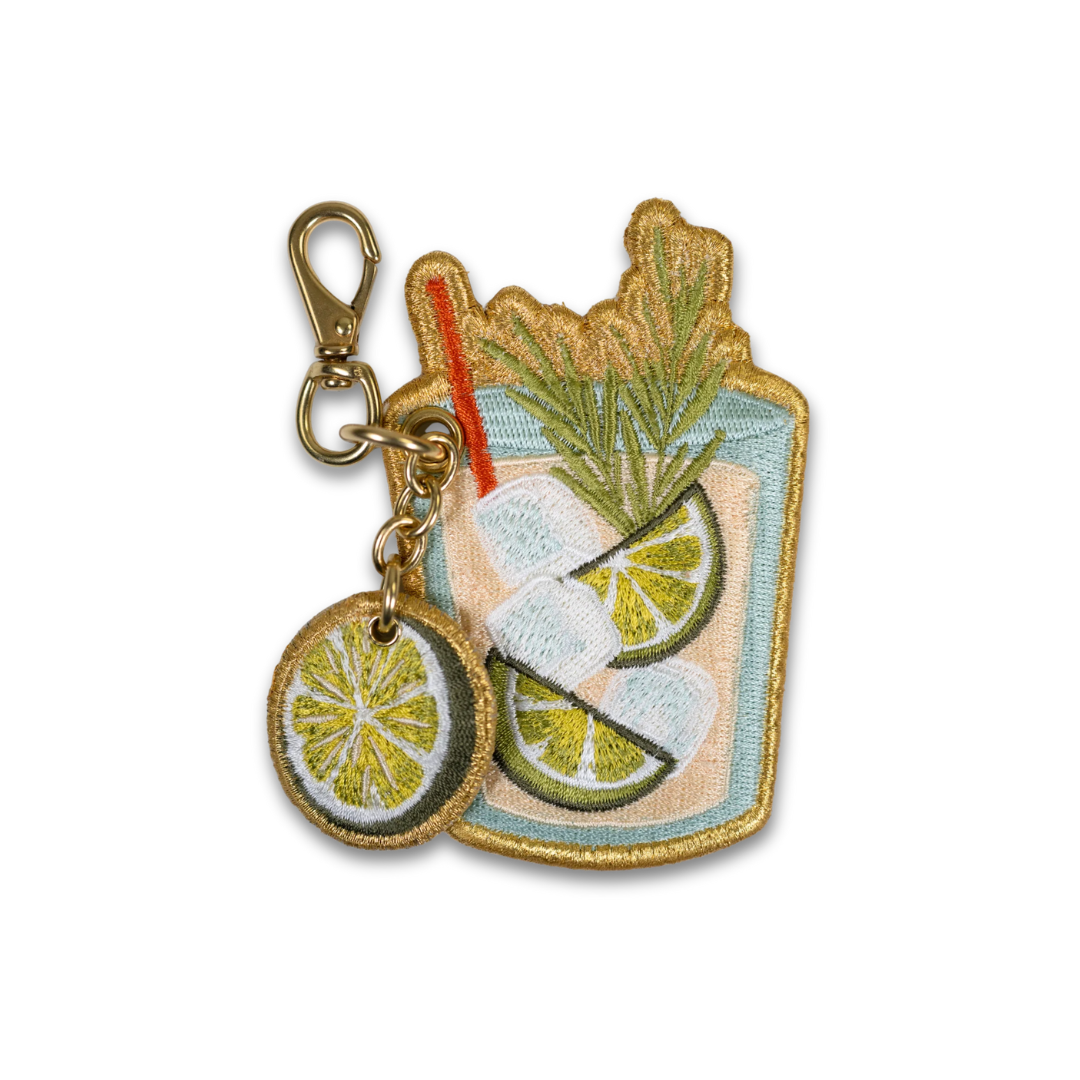 Embroidered cocktail keychain featuring a light blue margarita glass with lime slices, a red straw, and a round hanging charm of a lime wheel, attached to a gold-tone lobster clasp on a white background.